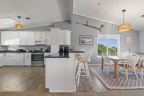 A modern kitchen with white cabinetry, a central island, and a dining area with white chairs. - Walk to Beach 5BR Waterview Dog Friendly | Pool (Duck)
