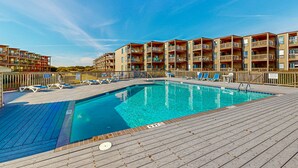 A swimming pool with lounge chairs and a wooden deck, surrounded by multi-story buildings with balconies. - 1BR Oceanfront condo with a pool and tennis court (Duck)