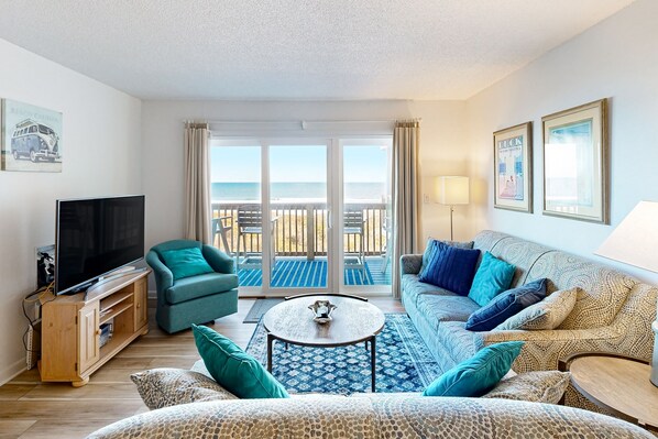 A living room with a TV, sofa, and coffee table, a balcony with a view of the sea, and a framed picture on the wall. - 1BR Oceanfront condo with a pool and tennis court (Duck)