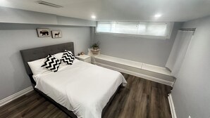 A bedroom with a bed, a nightstand, a bench, a window, and wall art. - Soft landing spot | Relaxed basement hideout (Ajax)