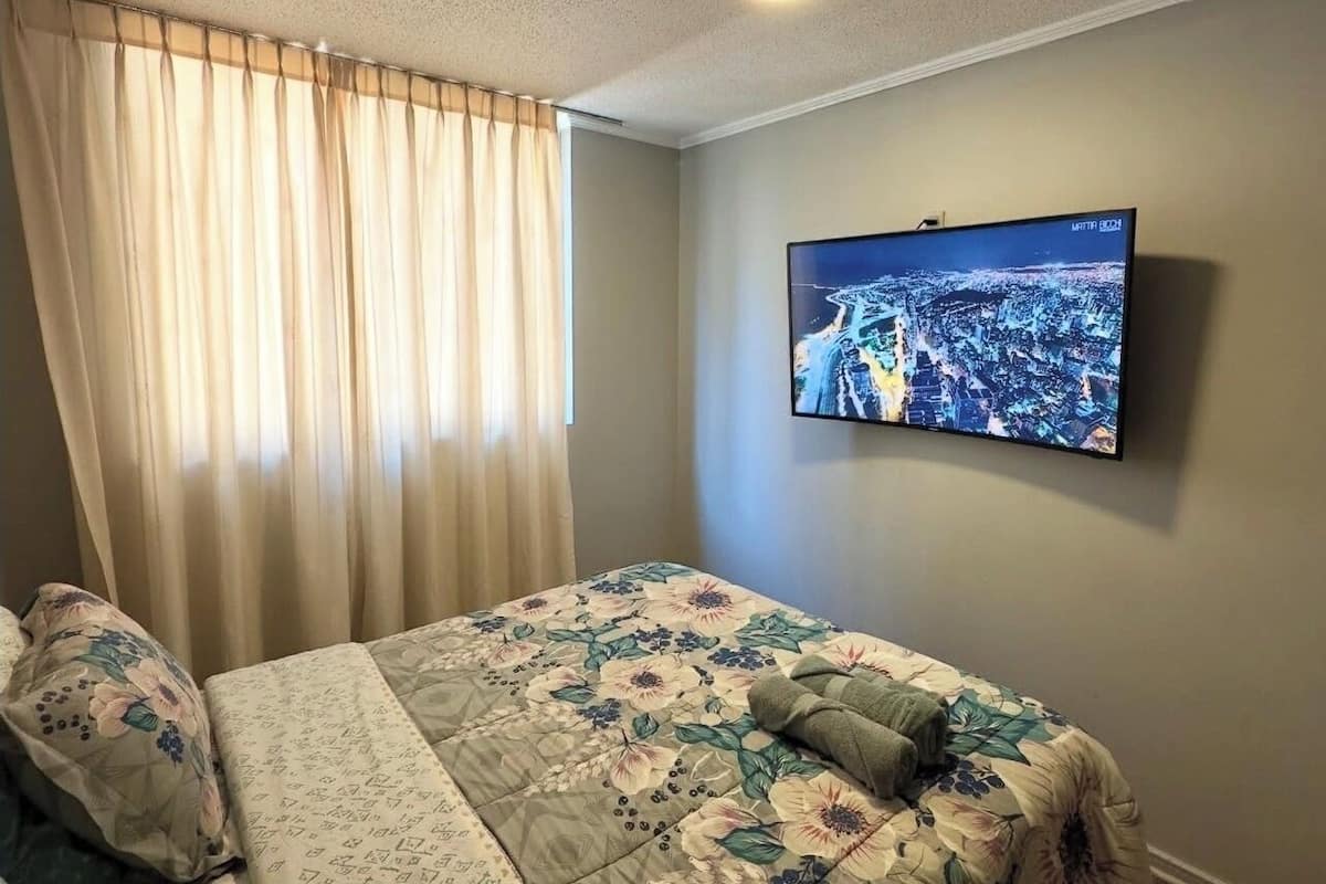 A bedroom with a bed, curtains, a television on the wall, and a ceiling light.