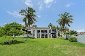 A modern two-story house with a large front lawn, palm trees, and a clear blue sky.