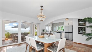 Una cocina moderna con armarios blancos, un comedor con mesa de vidrio y vista al océano a través de amplios ventanales correderos.