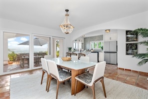 A modern kitchen with white cabinets, a dining area with a glass table, and a view of the ocean through large sliding doors.