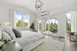 A bedroom with a large bed, a chandelier, a view of palm trees, and a sliding glass door to an outdoor area.
