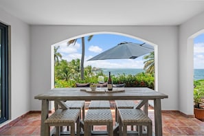 A table with woven chairs, a bottle, and glasses, set against a view of the ocean and palm trees.