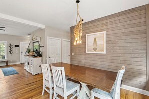 A dining area with a wooden table and white chairs, a hanging light fixture, and a framed picture on the wall. - KDS0900: BlueDream | Updated | Dog Friendly | Deck | Garage Parking (Kill Devil Hills)