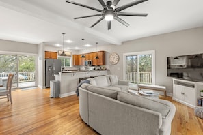 A spacious living room with a grey sofa, wooden flooring, a ceiling fan, and a kitchen area with wooden cabinets and a refrigerator. - KDS0900: BlueDream | Updated | Dog Friendly | Deck | Garage Parking (Kill Devil Hills)
