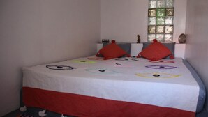 Una cama con una colcha blanca y roja adornada con ojos de colores, almohadas rojas y una ventana con marco verde.