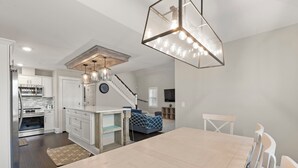 A modern kitchen with white cabinetry, a dining area with a wooden table and chairs, and a staircase leading to another level. - KDS0705: Beachy Dunes OBX | Newly-Built | Hot Tub | Deck | W/D (Kill Devil Hills)