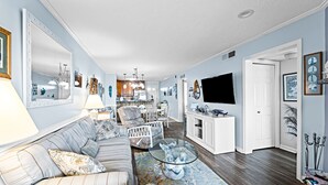 A spacious living room with a striped sofa, a glass coffee table, and a large mirror. - Serene 2BR Oceanfront 3rd-Floor | Balcony | Pool (Kill Devil Hills)