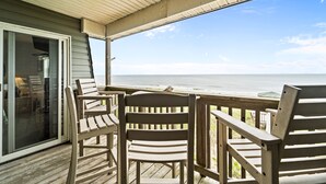 A wooden deck with two chairs facing a glass door leading to a room. - Serene 2BR Oceanfront 3rd-Floor | Balcony | Pool (Kill Devil Hills)