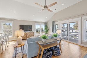 A spacious living area with a ceiling fan, large windows, a TV, and a dining table.