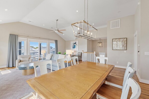 A spacious living room with a large wooden dining table, white chairs, a ceiling fan, and a view of the outside.