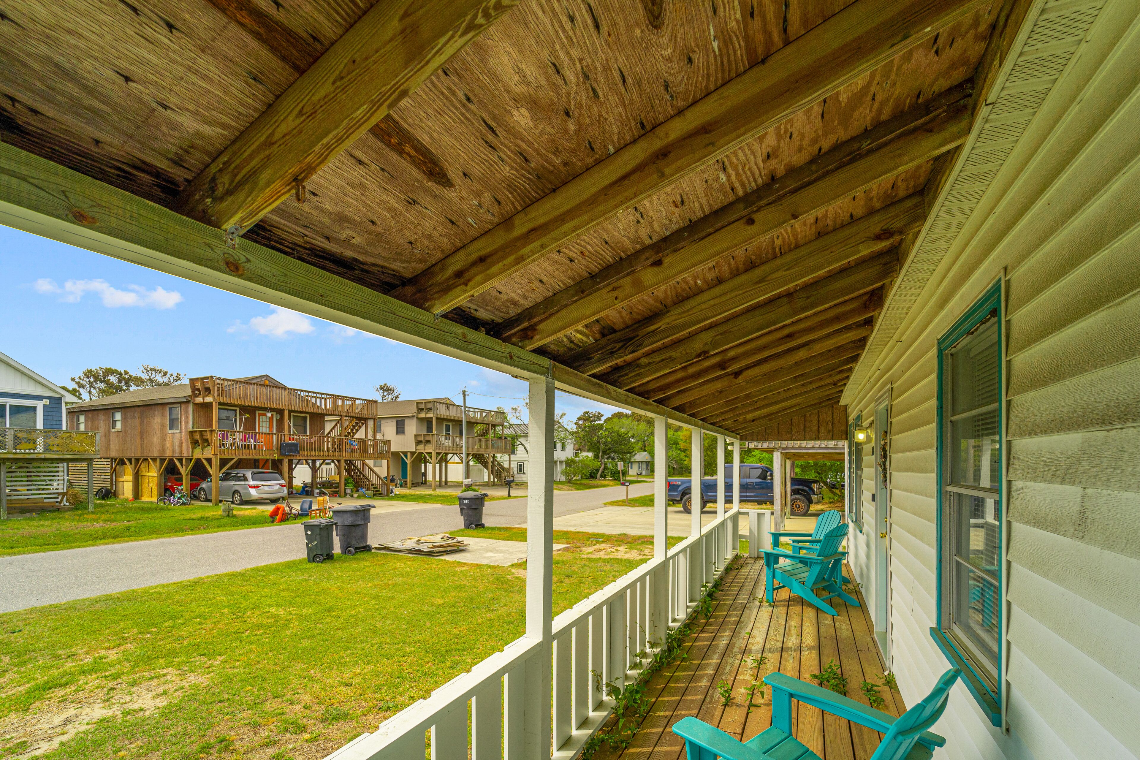 A covered porch with wooden beams and white railings, overlooking a street with houses and parked cars.