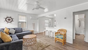 Amplia sala de estar con un sofá gris, una mesa de centro redonda de madera, una silla de mimbre y una zona de cocina con taburetes blancos.
