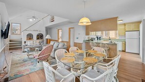 A spacious living area with a round glass table, wicker chairs, and a sofa. A kitchen with wooden cabinets and a dining area with a table and chairs are visible.