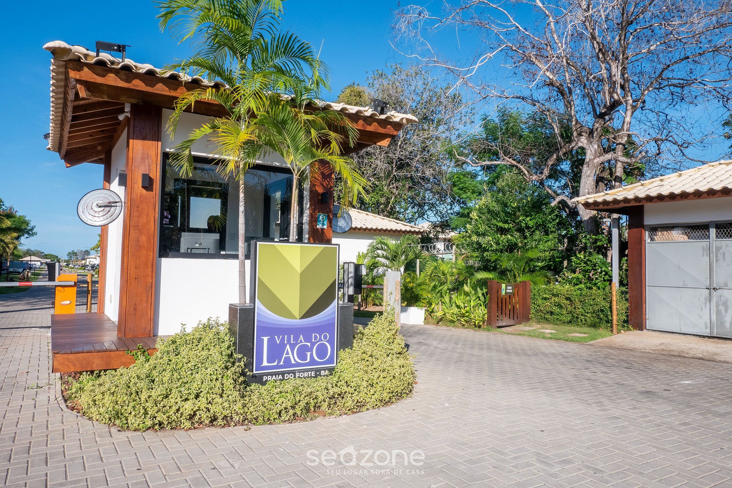 A modern villa with a wooden facade, a sign reading "Vila do Lago," and a paved entrance area with a small garden.