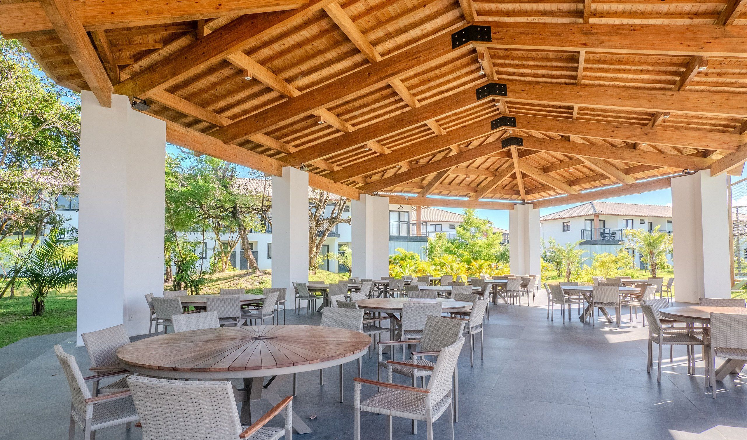 An outdoor patio area with round wooden tables and wicker chairs, surrounded by white pillars and a wooden ceiling structure.