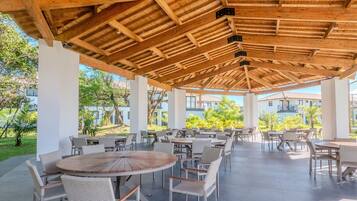 An outdoor patio area with round wooden tables and wicker chairs, surrounded by white pillars and a wooden ceiling structure.