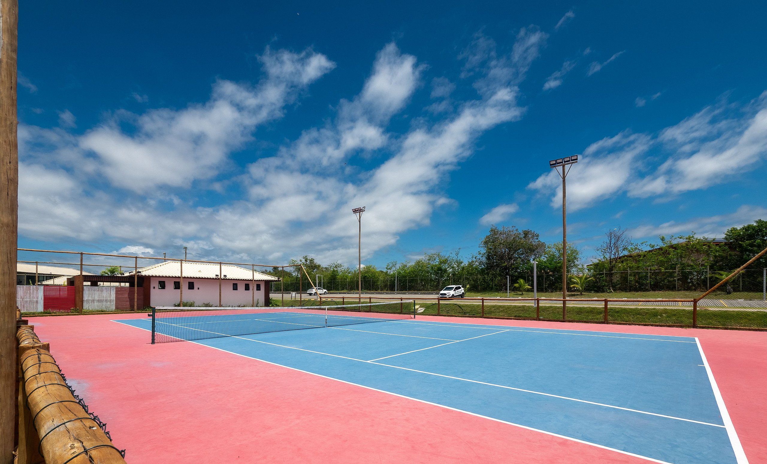 An outdoor tennis court with a net, surrounded by a fence and trees.