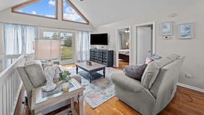 A spacious living room with a high ceiling, a ceiling fan, a large window, a flat-screen TV, a dark wooden cabinet, a grey sofa, a glass coffee table, and a patterned rug.