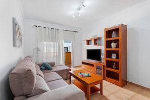 A living room with a sofa, TV, wooden cabinet, and a window with curtains. - Apartment at Playa San Juan (Playa San Juan)
