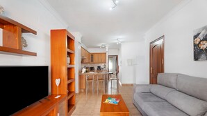 A living room with a TV, wooden shelves, a grey sofa, and a dining area in the background.