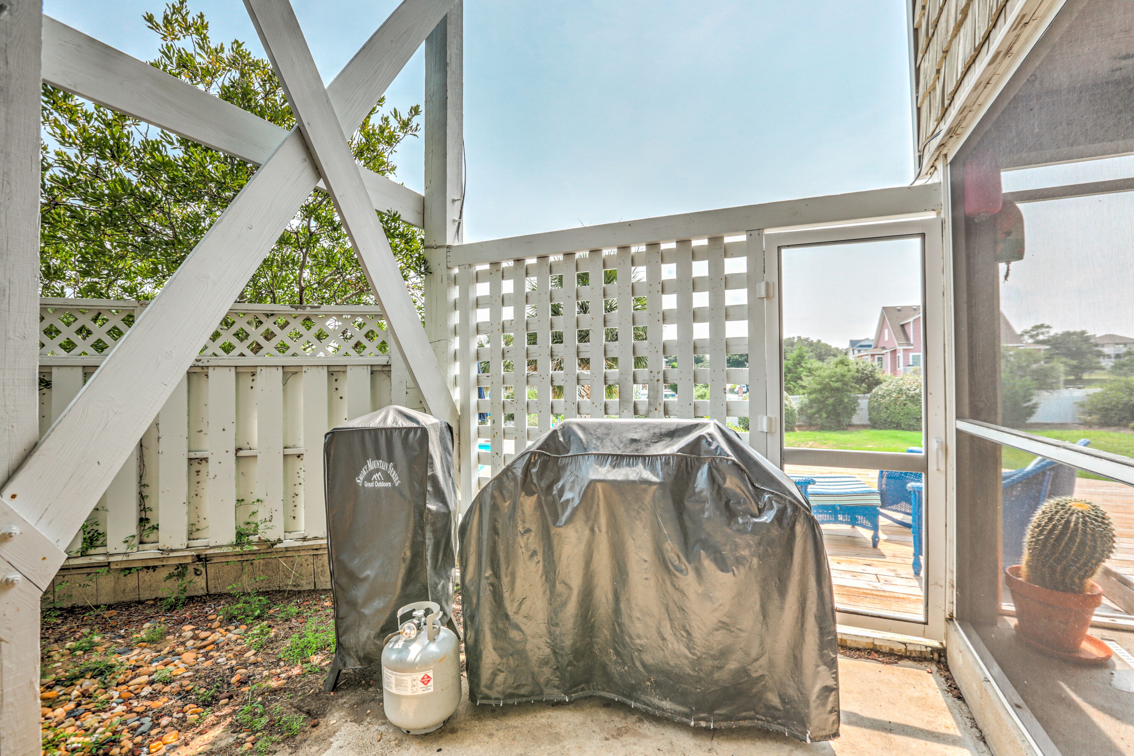 Un espace barbecue extérieur abrité, équipé d’un gril et d’une bouteille de propane, avec une clôture blanche et une vue sur une maison et de la verdure en arrière-plan.