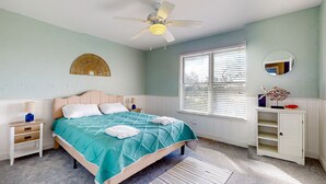 Une chambre à coucher comprenant un lit, des tables de chevet, un ventilateur de plafond, une fenêtre avec des stores, un miroir et un petit meuble.