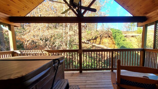 A wooden deck with a bench and a chair, a ceiling fan, and a view of trees and a rocky landscape. - Creekside - 1BR King Cabin (Gatlinburg)