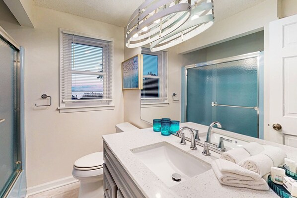 Un baño con un lavabo blanco, dos grifos, un espejo y un inodoro. Hay dos ventanas con persianas, una de ellas con un cuadro.