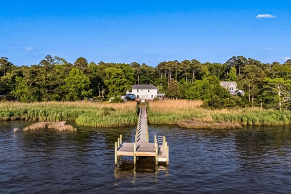 Une jetée en bois s’étendant sur une étendue d’eau calme, entourée d’une végétation luxuriante et de quelques maisons en arrière-plan.
