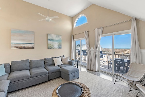 A spacious living room with a grey sectional sofa, a round coffee table, and a view of the beach through large windows.