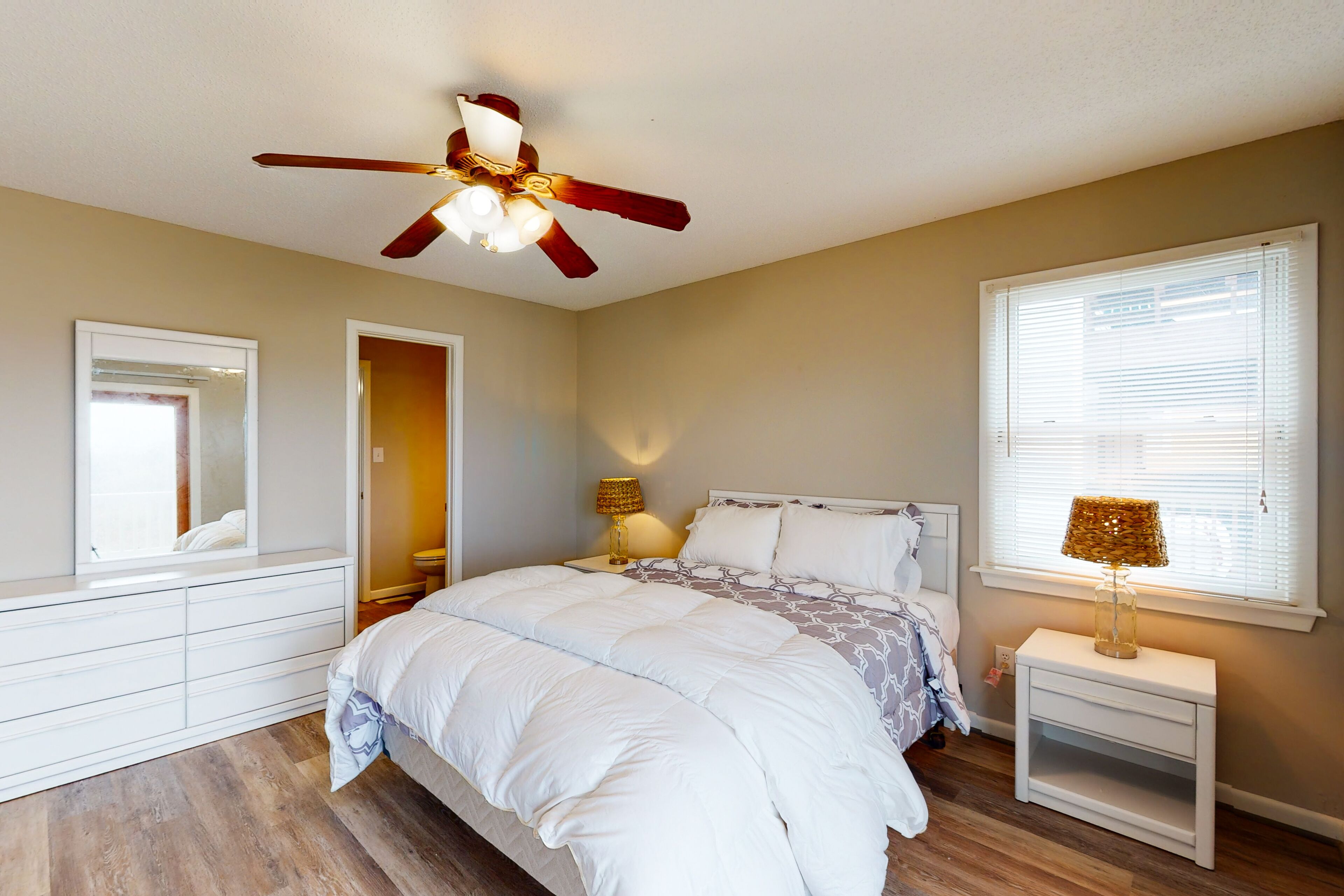 A bedroom with a bed, a ceiling fan, a nightstand with a lamp, a window with blinds, and a mirror.