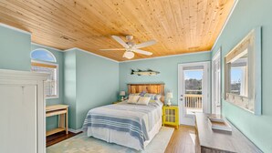 A bedroom with a wooden ceiling, a bed with a striped bedspread, a nightstand, a window with a view, and a fish decoration on the wall. - Refreshing 4BR Dog Friendly | Balcony | W/D (Nags Head)