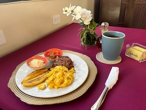 A breakfast plate with rice, beans, scrambled eggs, and plantains on a table with a vase of flowers, a cup, and a tissue box. - Hostal Boutique Isabel (Ticuantepe)