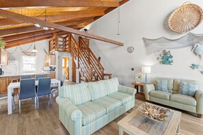 A spacious living area with a high ceiling, wooden staircase, a dining table with chairs, a striped sofa, a green sofa, and a decorative wooden bowl on a coffee table. - Large 3BR Oceanfront | Deck | Tennis | W/D (Nags Head)