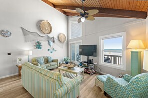 A living room with a TV, sofas, a coffee table, and a view of the ocean. - Large 3BR Oceanfront | Deck | Tennis | W/D (Nags Head)