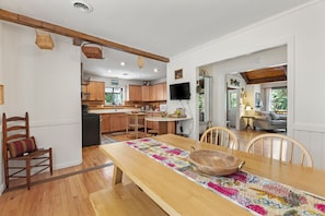 A kitchen with wooden cabinets, a dining table with a floral runner, and a TV mounted on the wall. - Tranquil 4BR Waterview | Fireplace | Deck (Southern Shores)
