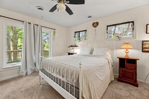 Un dormitorio con cama, mesita de noche, lámpara, ventilador de techo y ventanas con cortinas.