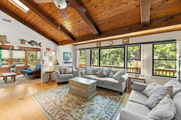 A spacious living room with wooden beams, large windows, a sofa, a coffee table, and a rug. - Tranquil 4BR Waterview | Fireplace | Deck (Southern Shores)
