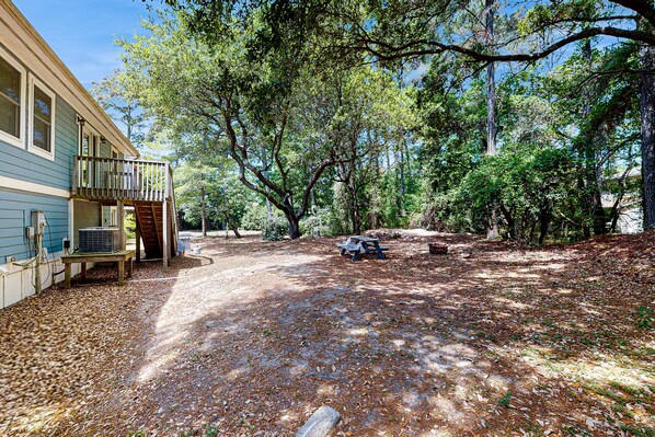 A house with a covered porch and a wooden deck, surrounded by trees and a gravel driveway. - KDN4512: Sandy Feet Retreat | Dog Friendly | Fireplace | Theater Room (Kill Devil Hills)