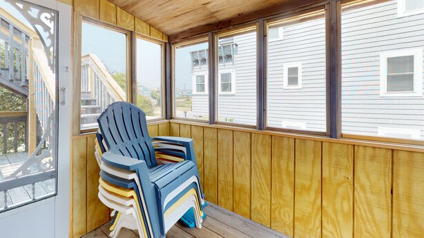 A covered porch with wooden walls and large windows, a blue chair, and a view of neighboring houses.