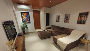 A modern living room with a brown sofa, a coffee table, and a TV on a stand. There are several paintings on the wall and a potted plant.