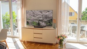 Una sala de estar con un televisor sobre un mueble bajo, un jarrón con flores y vistas al exterior.