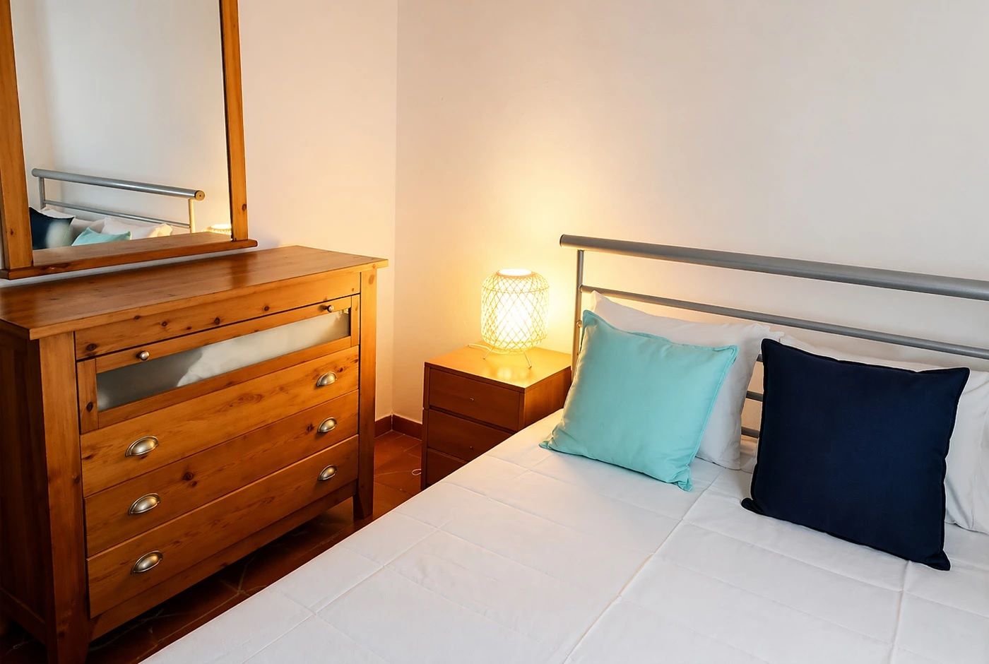 A bedroom with a bed, a wooden dresser with drawers, a bedside table, and a lamp.
