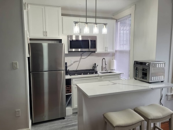 Una cocina moderna con un refrigerador de acero inoxidable, un microondas y un horno empotrado. Hay una isla blanca con taburetes de bar e iluminación colgante arriba.