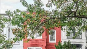 Un edificio de ladrillo rojo con adornos blancos, varias ventanas y un pequeño jardín delantero con un árbol.