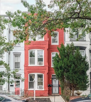 Un edificio de ladrillo rojo con adornos blancos, varias ventanas y un pequeño jardín delantero con un árbol.